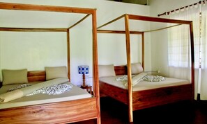 Dos camas con dosel de madera, con ropa de cama y almohadas color beige, una mesita con una lámpara de estampado azul y blanco, y una ventana con cortinas blancas.