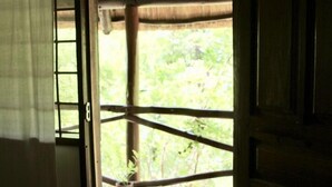 Une porte ouverte donnant sur un balcon doté d’un toit de chaume, d’un sol en béton et offrant une vue sur la verdure.