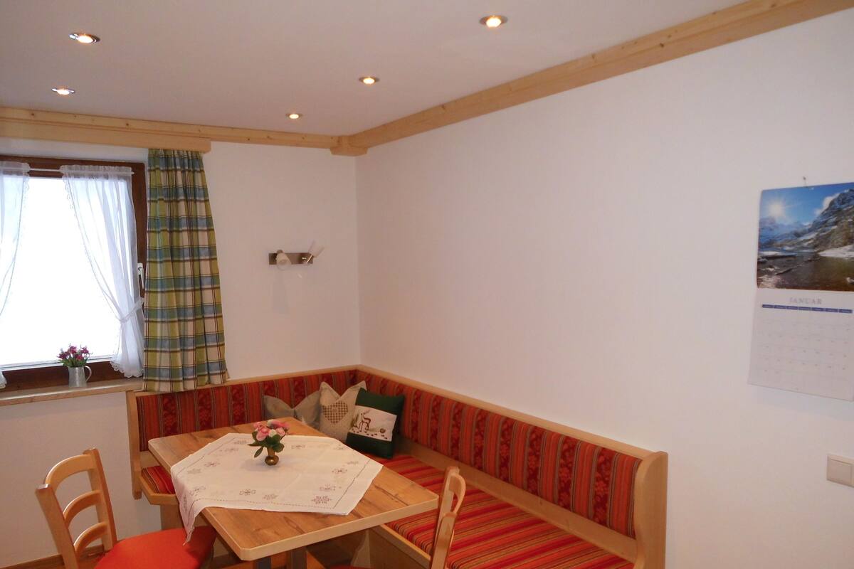 A cozy room with a wooden table, red cushioned chairs, a bench with striped cushions, a window with curtains, and a calendar on the wall.