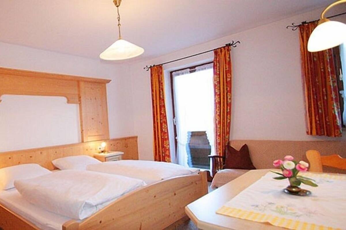 A hotel room with two single beds, wooden headboard, bedside tables with lamps, a window with curtains, and a table with a vase of flowers.