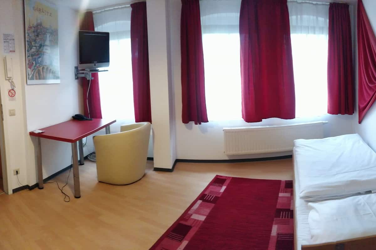 A hotel room with a bed, a desk, a chair, a television, and a window with red curtains.