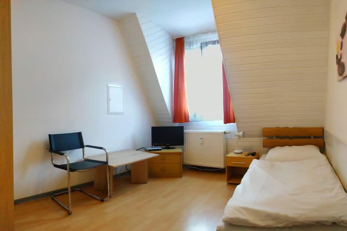 A small, well-lit room with a bed, a desk with a chair, a television, and a window with curtains.