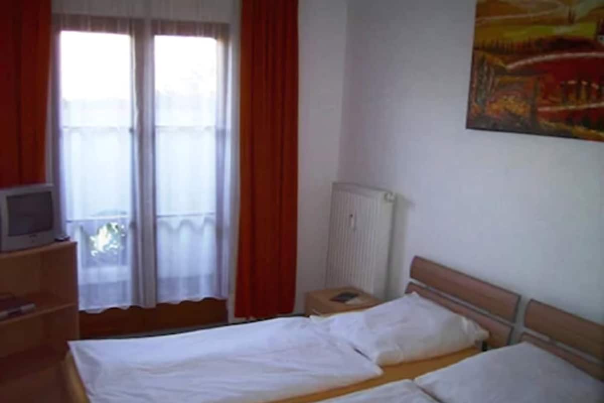 A room with two beds, a radiator, a television on a wooden stand, and a window with curtains.