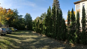 A paved courtyard with parked cars, tall evergreen trees, and a building with large windows. - Picobello Pension (Görlitz)