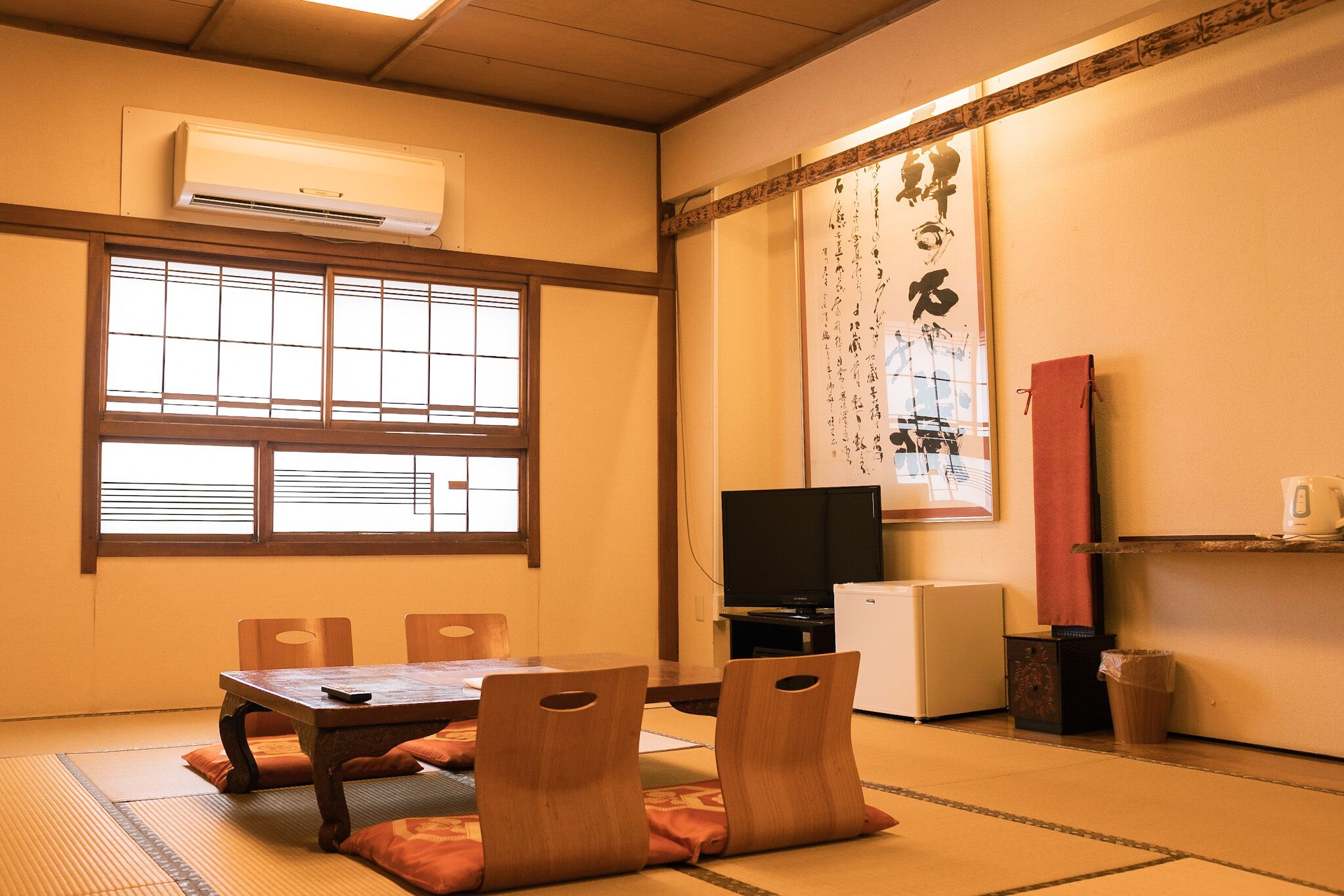 Une pièce de style japonais traditionnel, avec un sol recouvert de tatamis, une table basse, des chaises en bois, un téléviseur et un petit réfrigérateur.