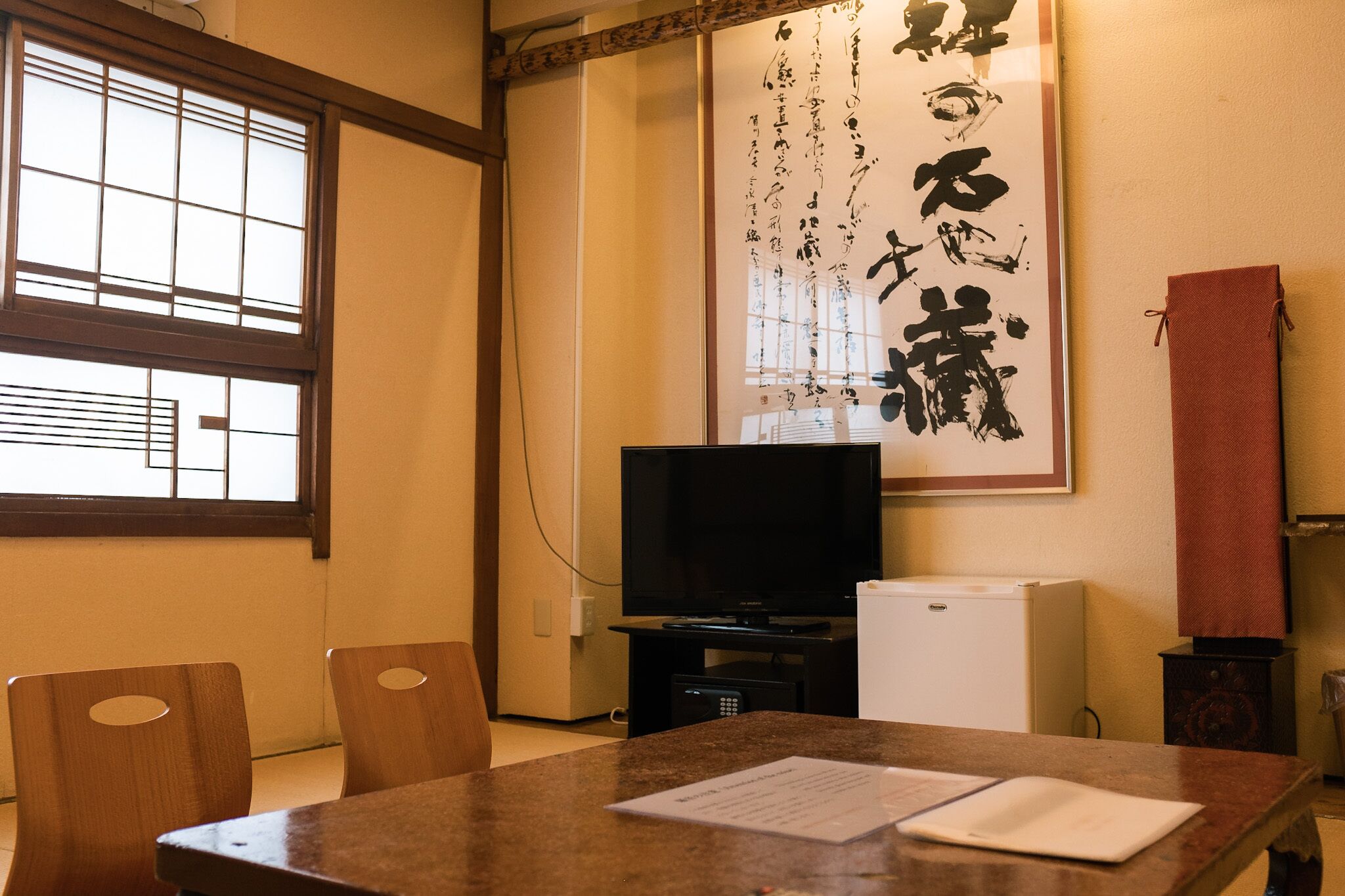 A room with a wooden table, chairs, a television, a small refrigerator, and a large calligraphy artwork on the wall.