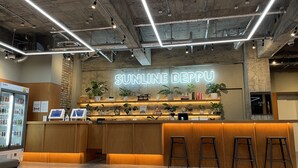 A modern bar area with a neon sign reading "SUNLINE BEPPU," wooden counters, bar stools, and a variety of plants and books on shelves.