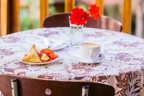 A table with a floral tablecloth, a plate of food, a cup of coffee, and a vase with red flowers.