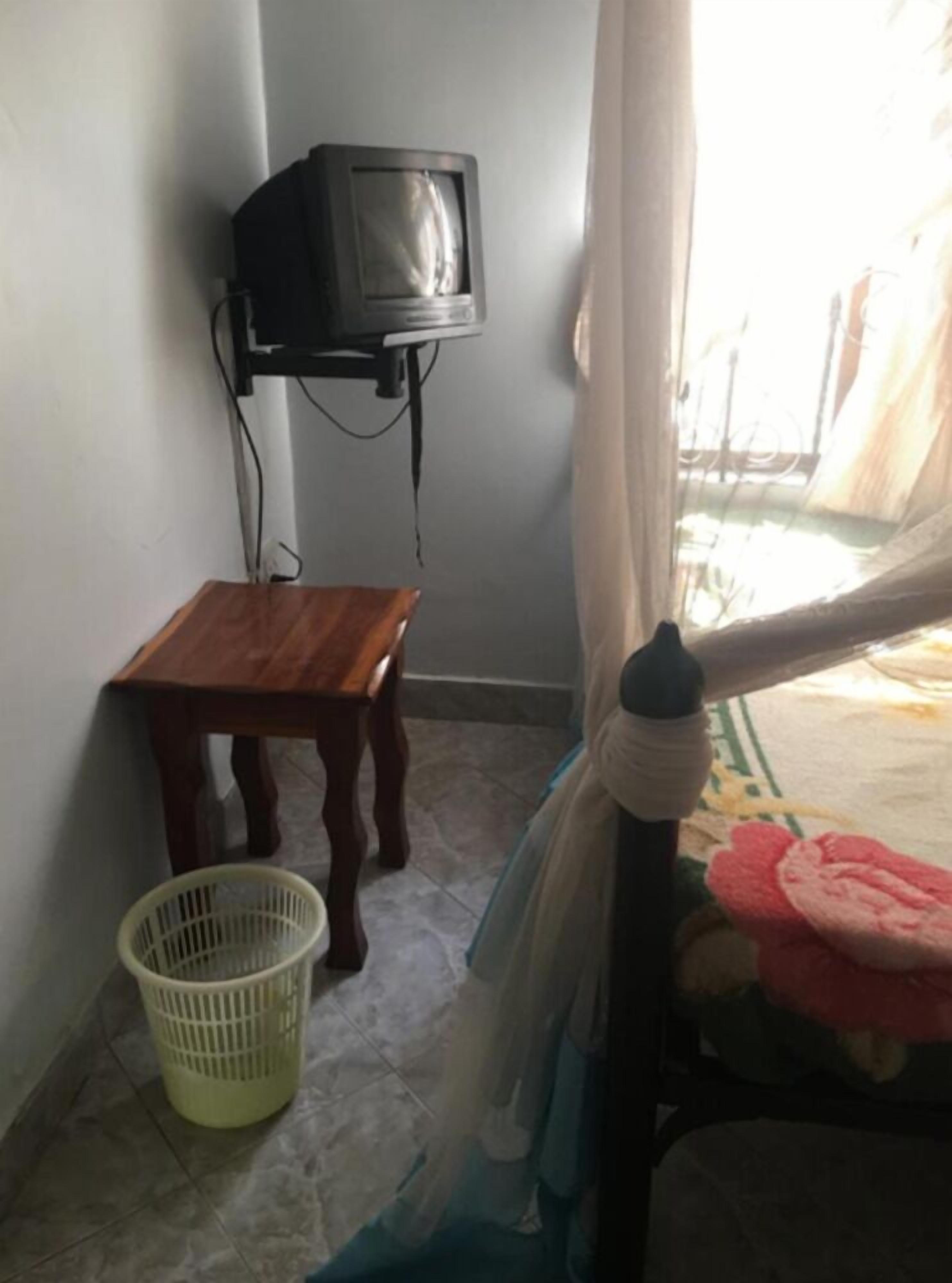 A room with a small wooden table, a television mounted on the wall, a plastic basket, and a bed with a floral bedspread.