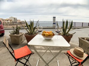 Une table avec un saladier de fruits et deux verres à vin, entourée de plantes en pot et de chaises d’extérieur.