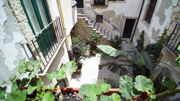 A narrow courtyard with a staircase leading to a second floor, surrounded by buildings with peeling paint and green shutters. - Casa Levante (Syracuse)