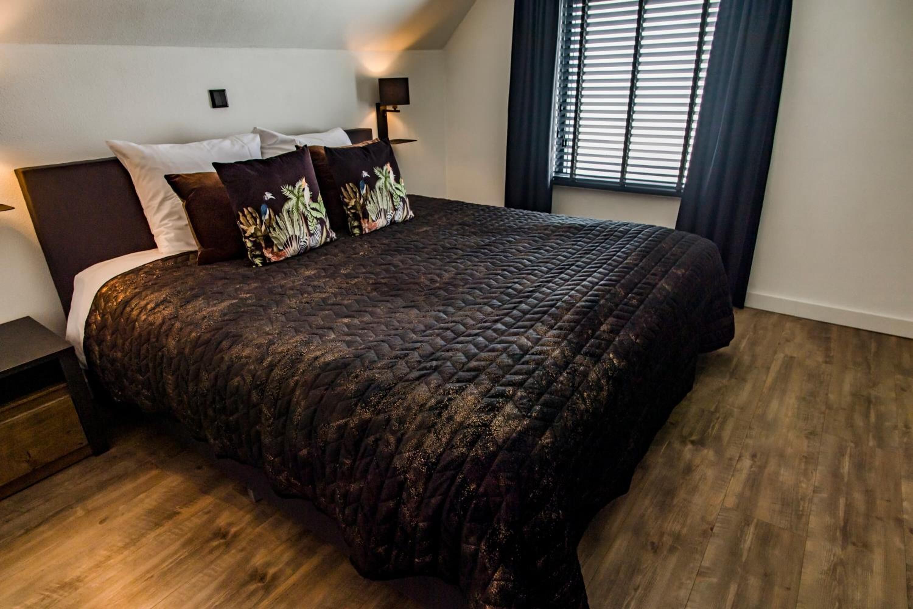 A bedroom with a large bed, dark bedding, and a window with blinds.