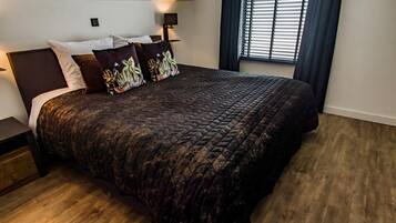 A bedroom with a large bed, dark bedding, and a window with blinds.