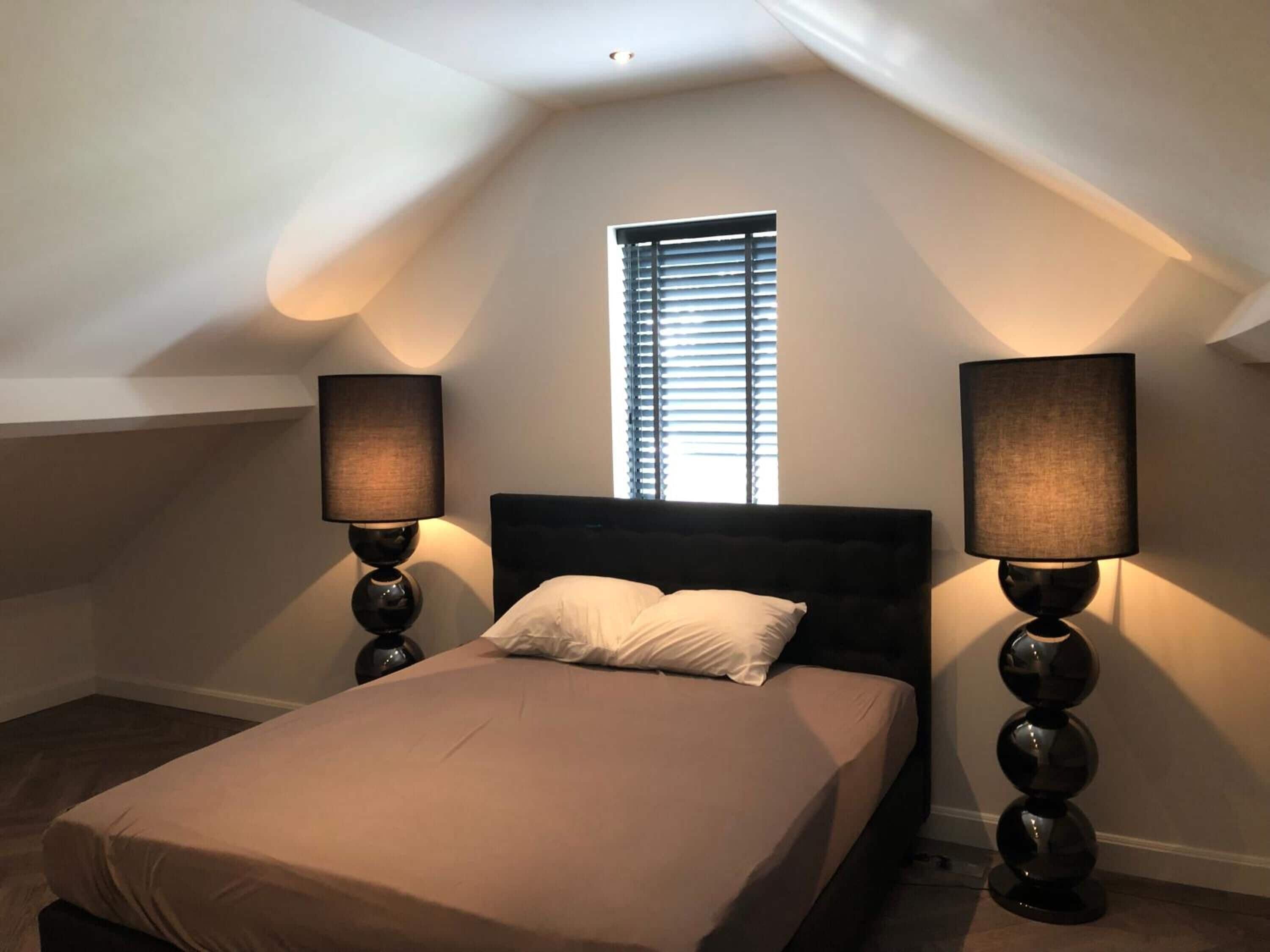 Een slaapkamer met een bed, twee bedlampen, een raam met zonwering en een schuine plafond.