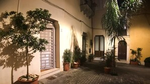 Um pátio com plantas em vasos, piso de azulejos e entrada de edifício com porta e janela.