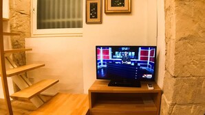 Quarto pequeno com uma estante de madeira onde está um aparelho de televisão exibindo um noticiário. Há duas fotos emolduradas na parede acima da TV.
