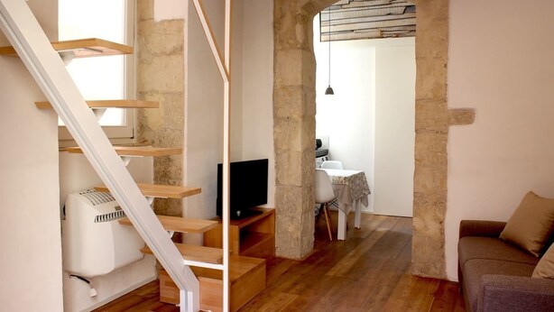 Uma escada de madeira com corrimão branco que leva a um quarto com televisão, uma mesa pequena e um sofá.