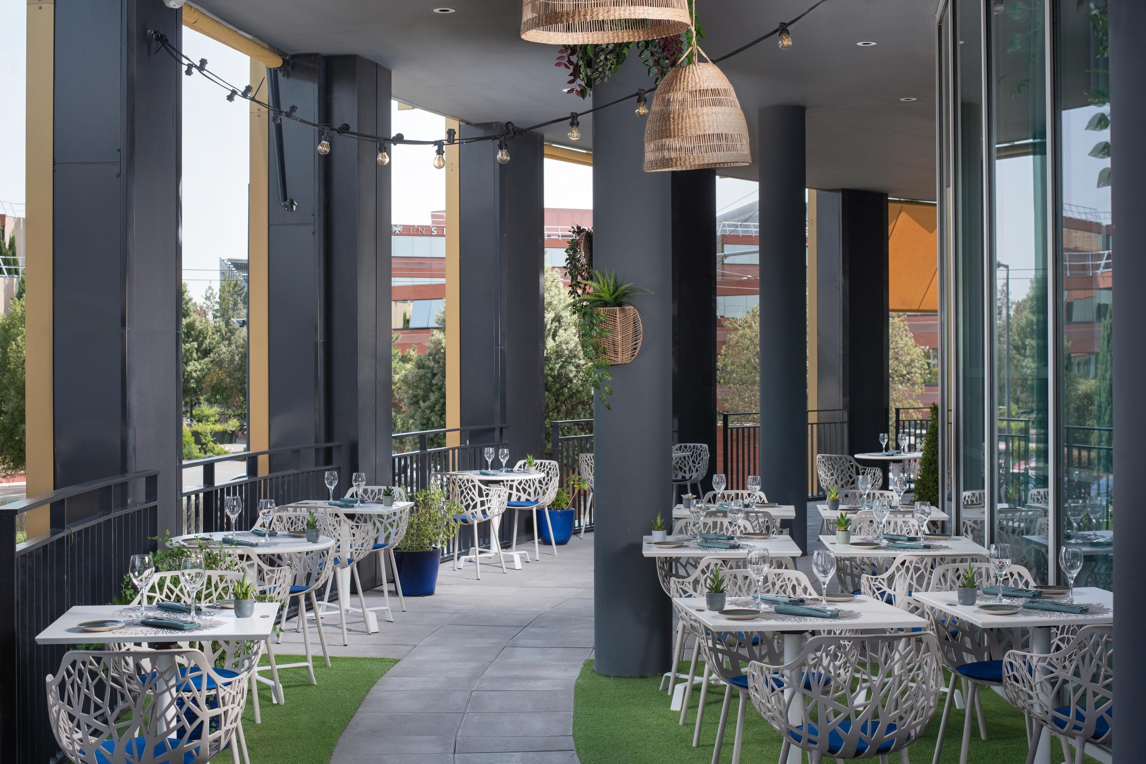 Un área de comedor al aire libre con mesas y sillas blancas, rodeada de amplias ventanas y columnas.