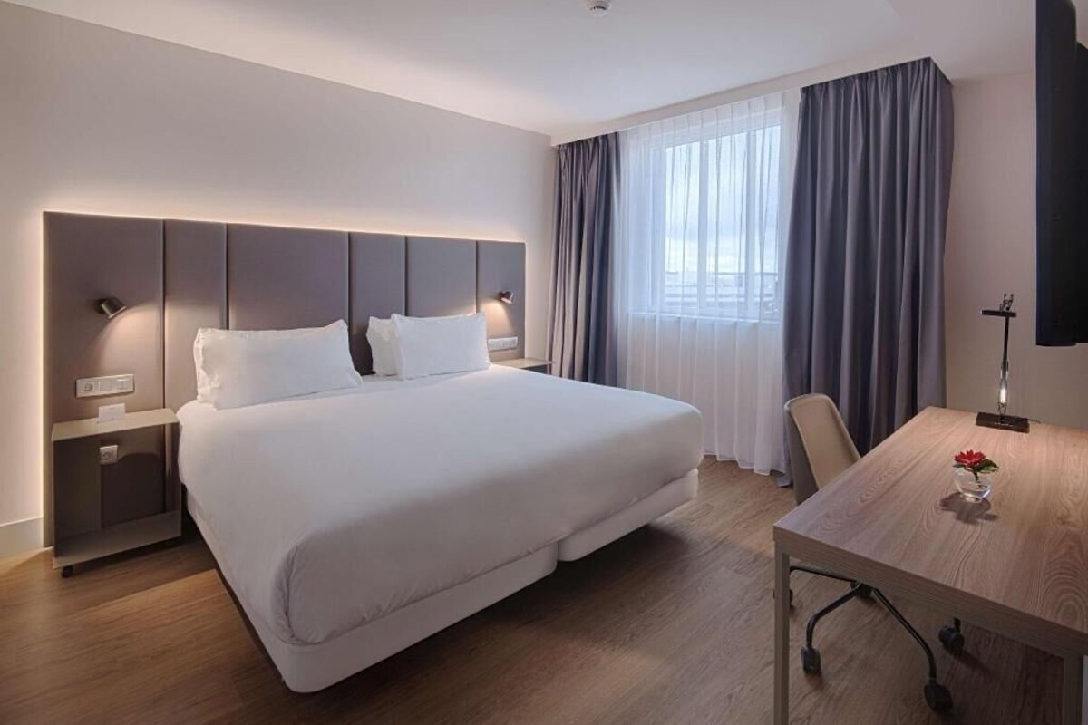 A hotel room with a large bed, a desk, a chair, a TV, and a window with curtains.