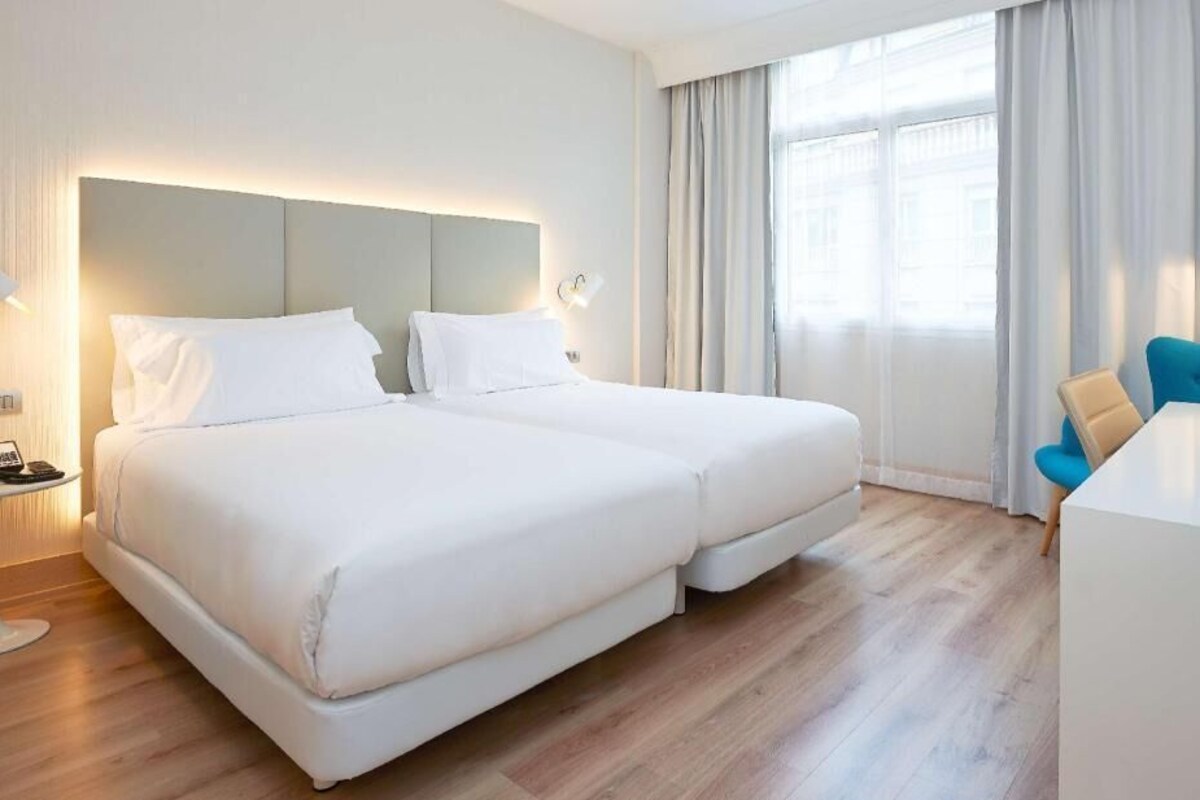 A hotel room with a double bed, a desk with a chair, a lamp, and a window with curtains.