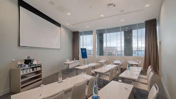 A conference room with tables, chairs, a projector screen, a coffee station, and a view of the city through large windows.