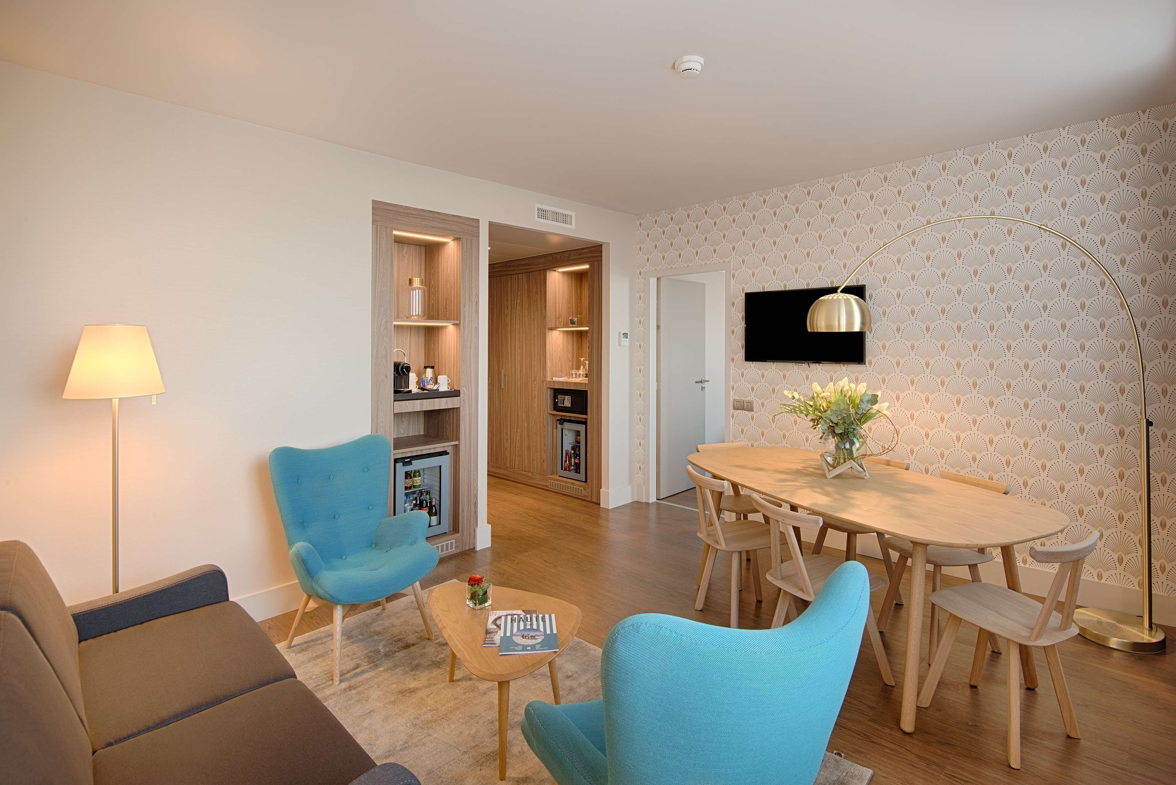 A modern living room with a sofa, two blue armchairs, a coffee table, and a dining area with a wooden table and chairs.