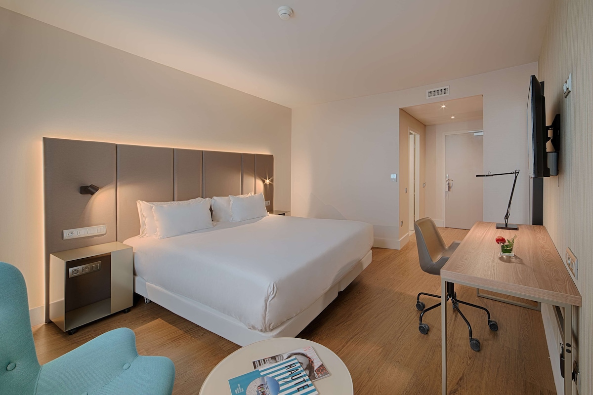 A modern hotel room with a large bed, a desk, a chair, and a small table with magazines.