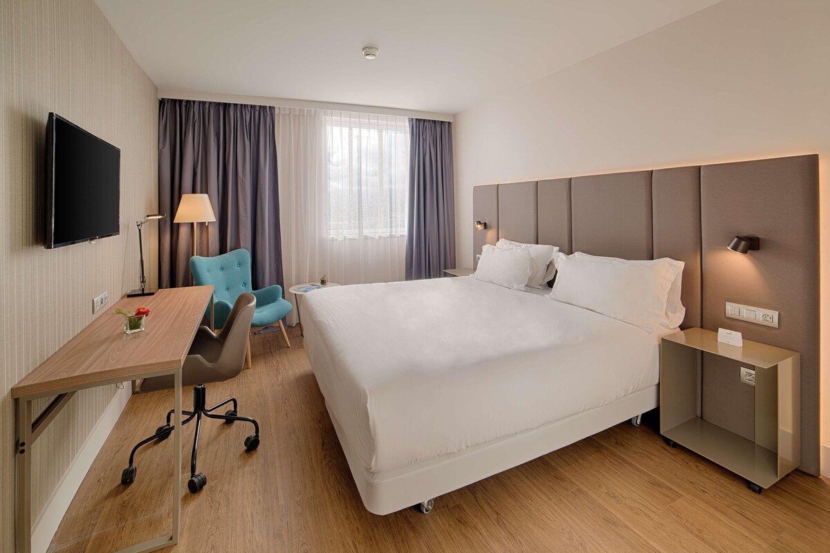 A hotel room with a bed, a desk, a chair, a TV, and a lamp.