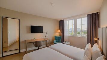 Habitación de hotel con cama, escritorio, silla, televisor, lámpara y ventana con cortinas.