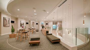 Een moderne hotellobby met een gebogen plafond, een zithoek met een bank en stoelen, een eettafel en een trap met glazen balustrades.