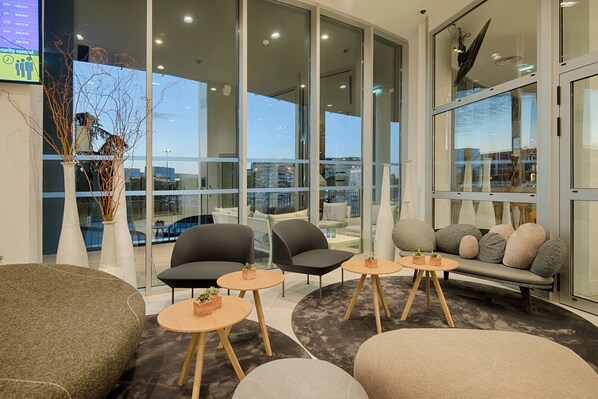 A modern lounge area with seating arrangements, including sofas and chairs, and a view of the cityscape through large windows. - NH Toulouse Airport (Blagnac)