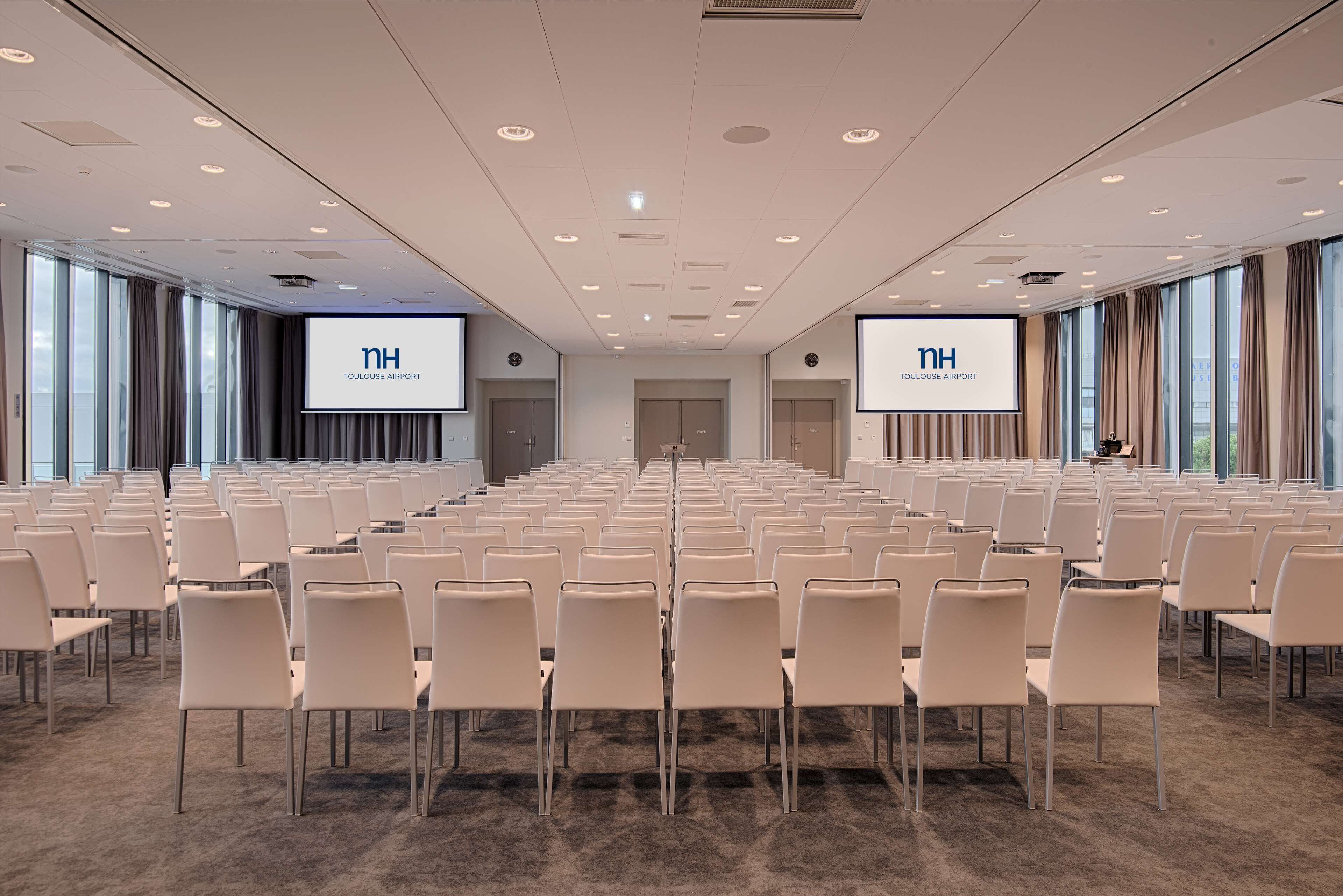 Un salón de conferencias con filas de sillas, dos pantallas grandes mostrando 'TH TOULOUSE AIRPORT' y suelo alfombrado.