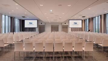 Un salón de conferencias con filas de sillas, dos pantallas grandes mostrando 'TH TOULOUSE AIRPORT' y suelo alfombrado.