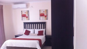 A neatly made bed with a white and red bedspread, a wooden headboard, and two red pillows. There are two paintings on the wall above the bed, and a black nightstand beside it. - Lediba Guest lodge (Maun)