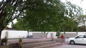 A paved outdoor area with steps leading to a pool, surrounded by trees and a white wall. - Lediba Guest lodge (Maun)