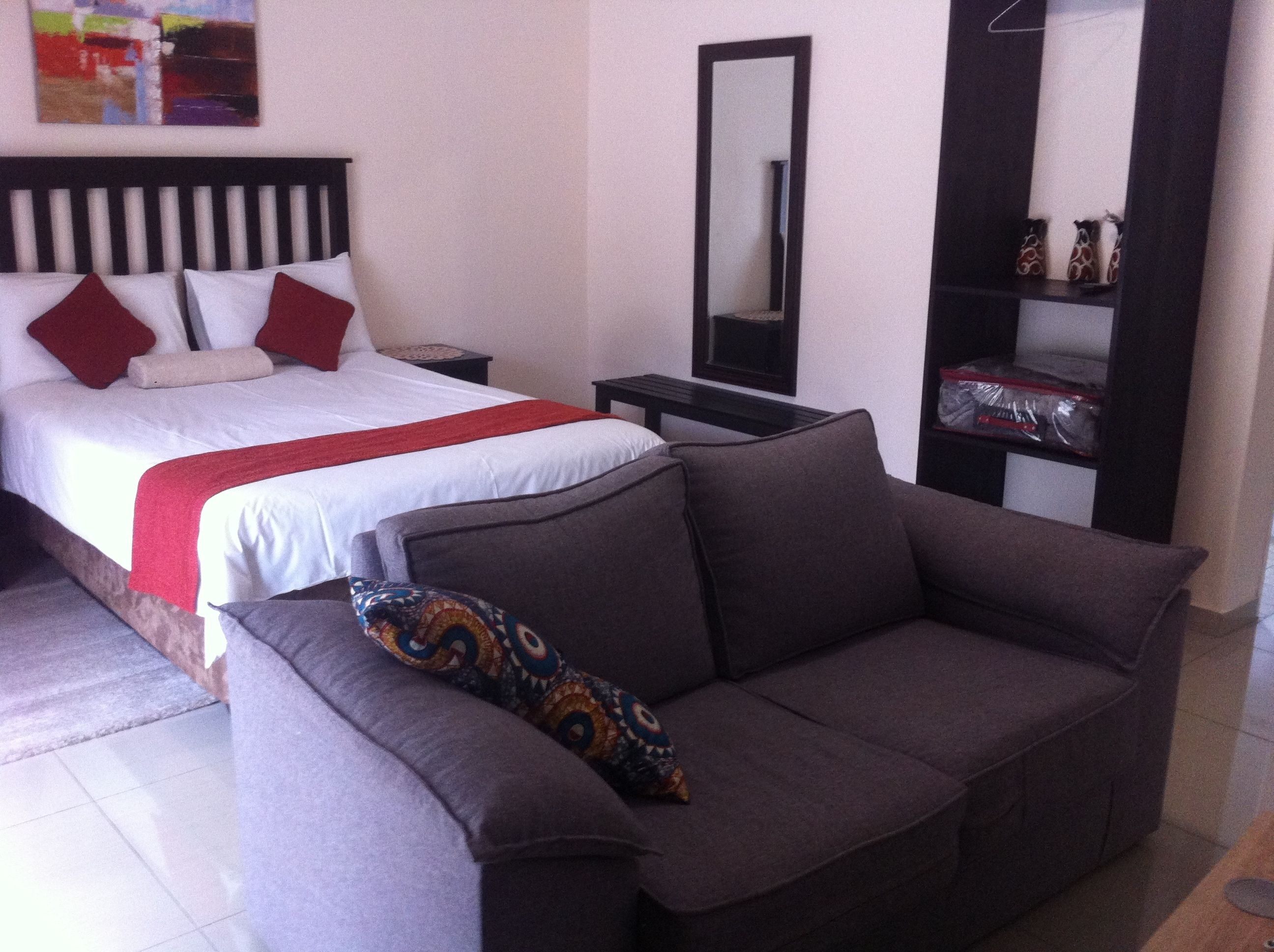 A hotel room with a bed, a sofa, a nightstand, a mirror, and a shelf with decorative items.