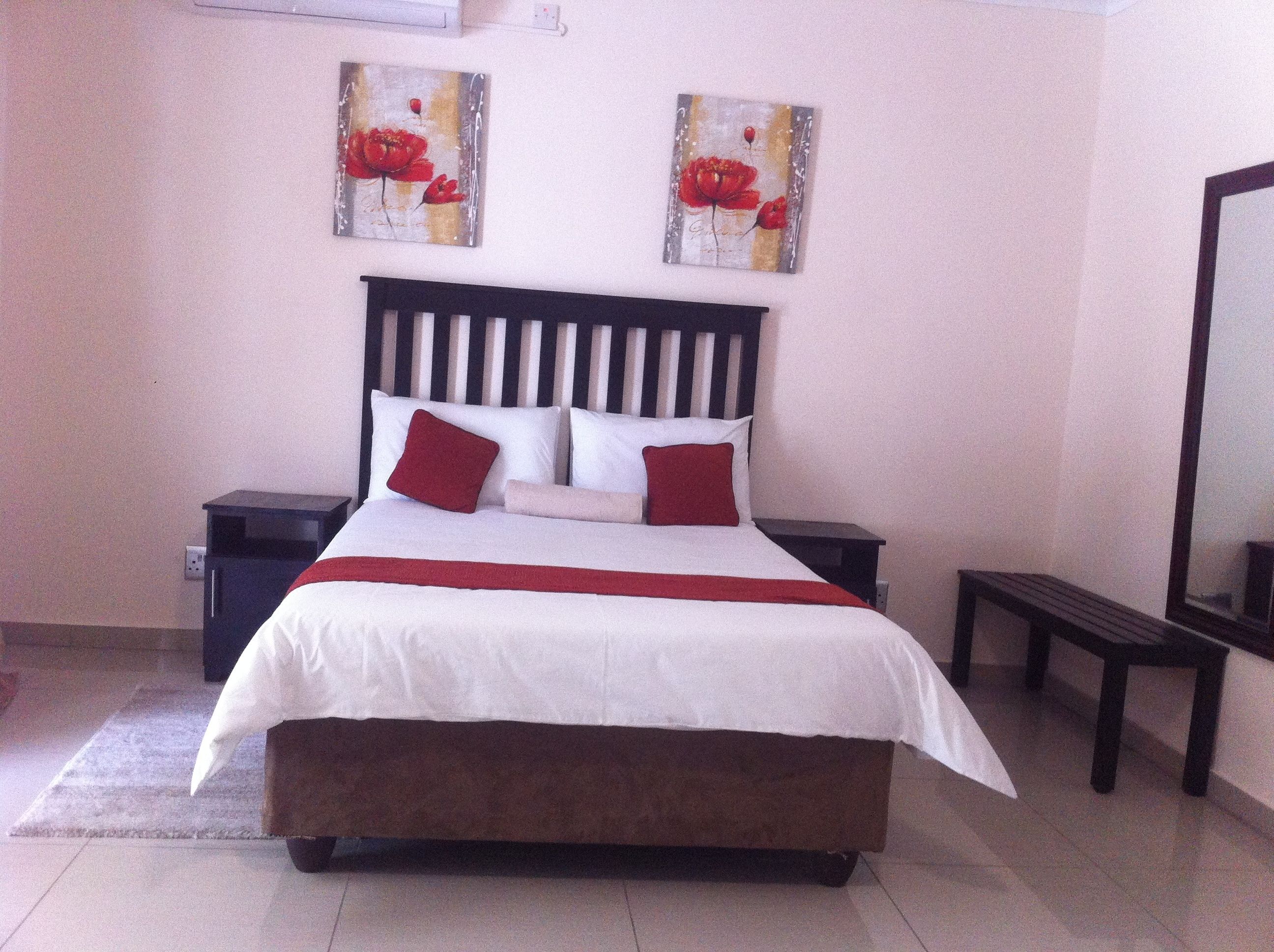 A neatly made bed with a white and red bedspread, two red pillows, and a wooden headboard. There are two paintings of red flowers on the wall above the bed.