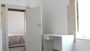 Une pièce avec un bureau blanc, un lit recouvert d’une literie à motifs floraux et une porte entrouverte donnant sur une autre pièce.