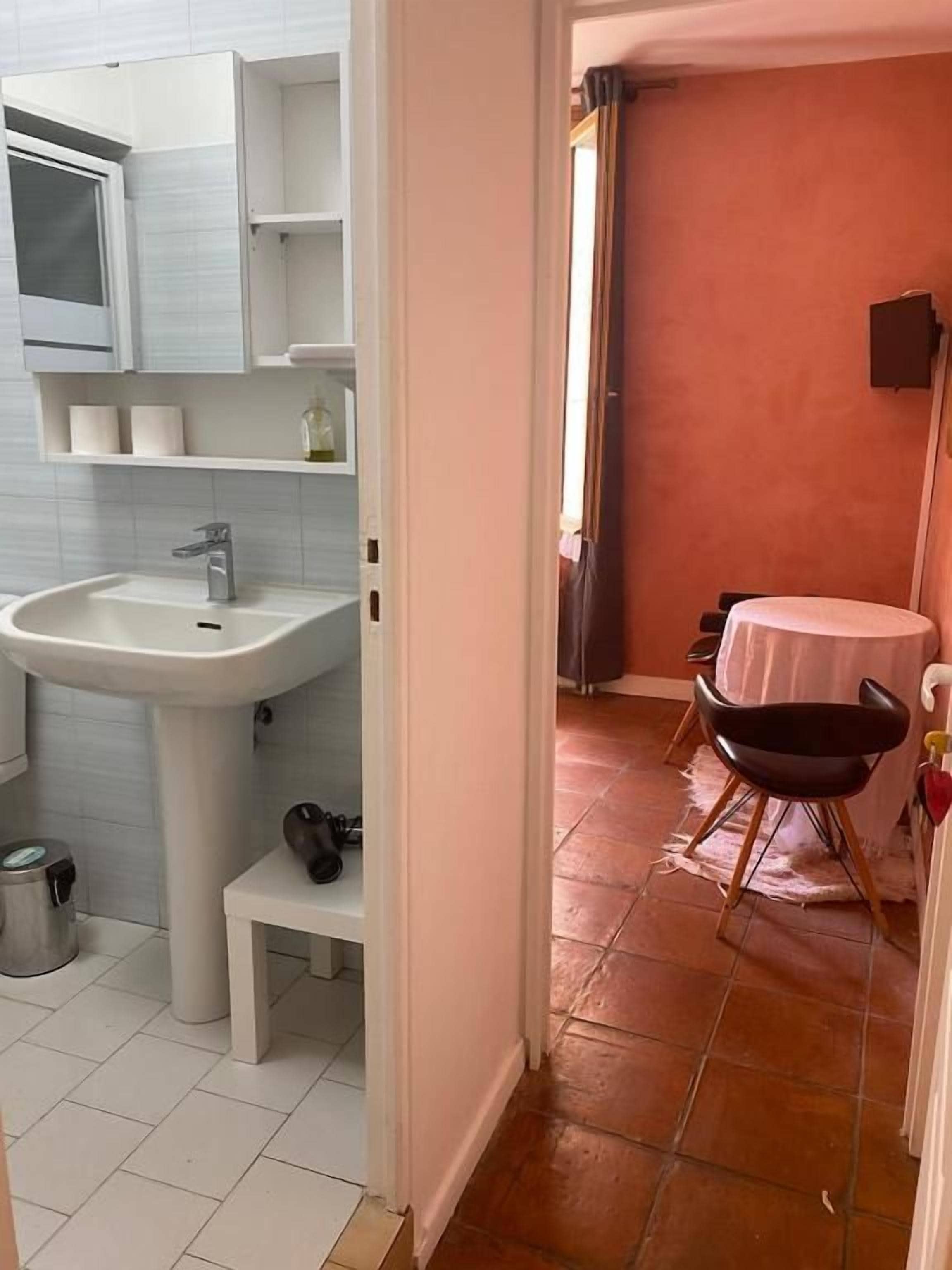 Een badkamer met een wastafel, spiegel en planken; een kleine tafel met stoelen; en een deur naar een kamer met een rode muur.