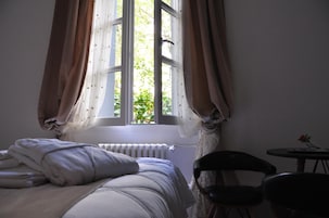 Un dormitorio con una cama, una silla, una mesa con una rosa y una ventana con cortinas.
