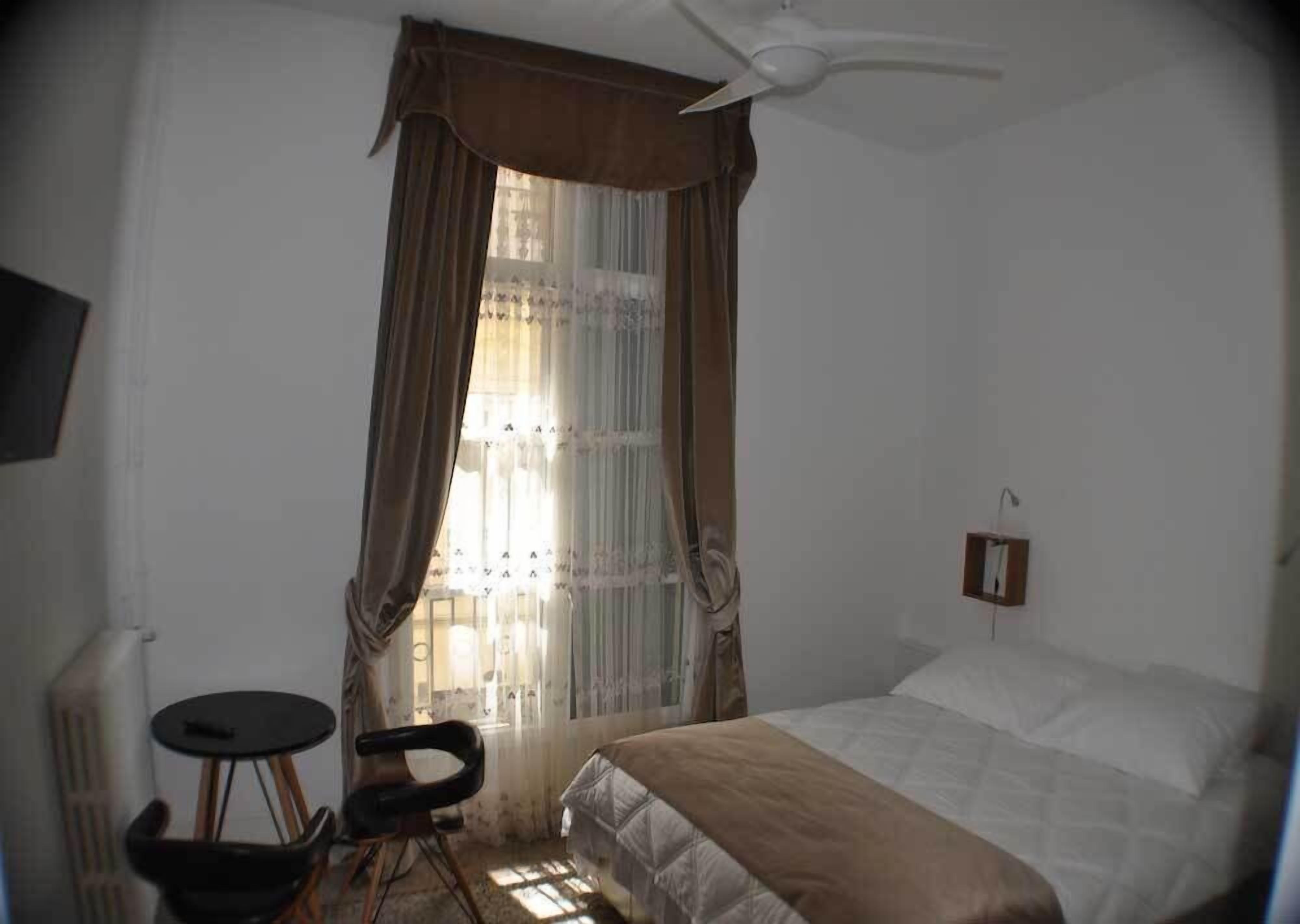 Une chambre à coucher comprenant un lit, une chaise, une petite table, un ventilateur de plafond et une fenêtre avec des rideaux.