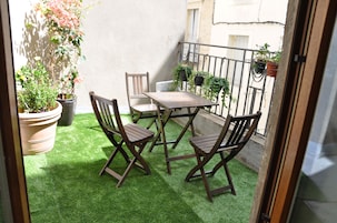 Un pequeño patio al aire libre con una mesa y sillas de madera, rodeado de plantas en macetas y un piso de césped artificial verde.