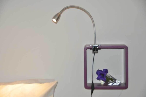 Espejo de marco cuadrado con una lámpara curva sobre él, reflejando una flor morada en un recipiente de vidrio.