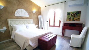 Un dormitorio con una cama, un reposapiés rojo, una silla blanca, un escritorio y un cuadro en la pared.
