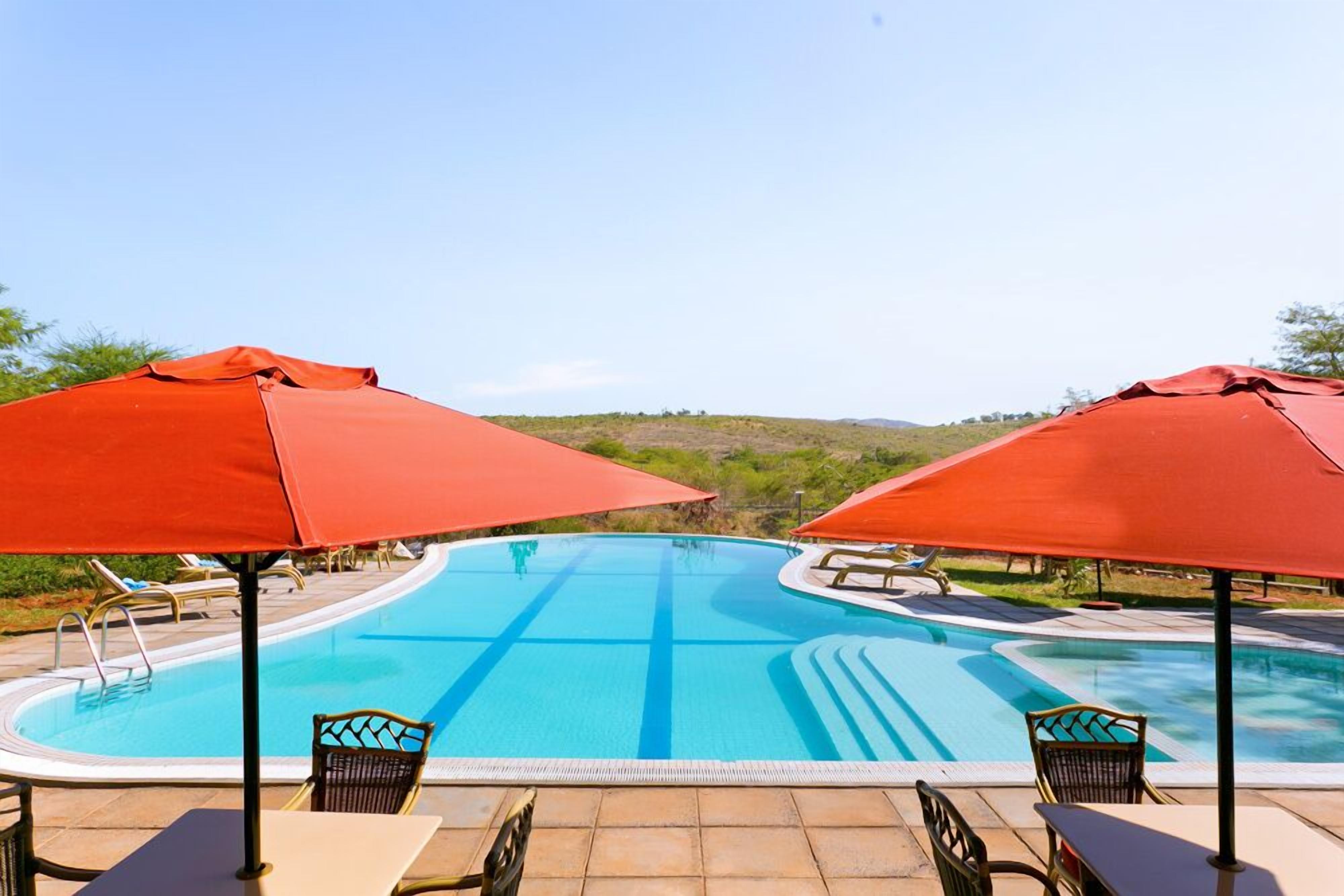 Una piscina con dos sombrillas rojas, sillas de descanso y vistas a colinas al fondo.
