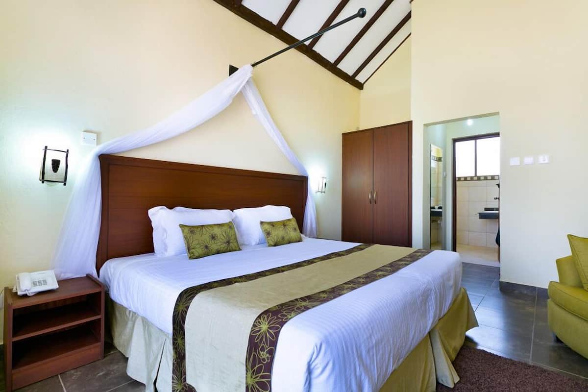 A hotel room with a large bed, a wooden headboard, a bedside table with a phone, a green chair, and a bathroom visible through an open door.