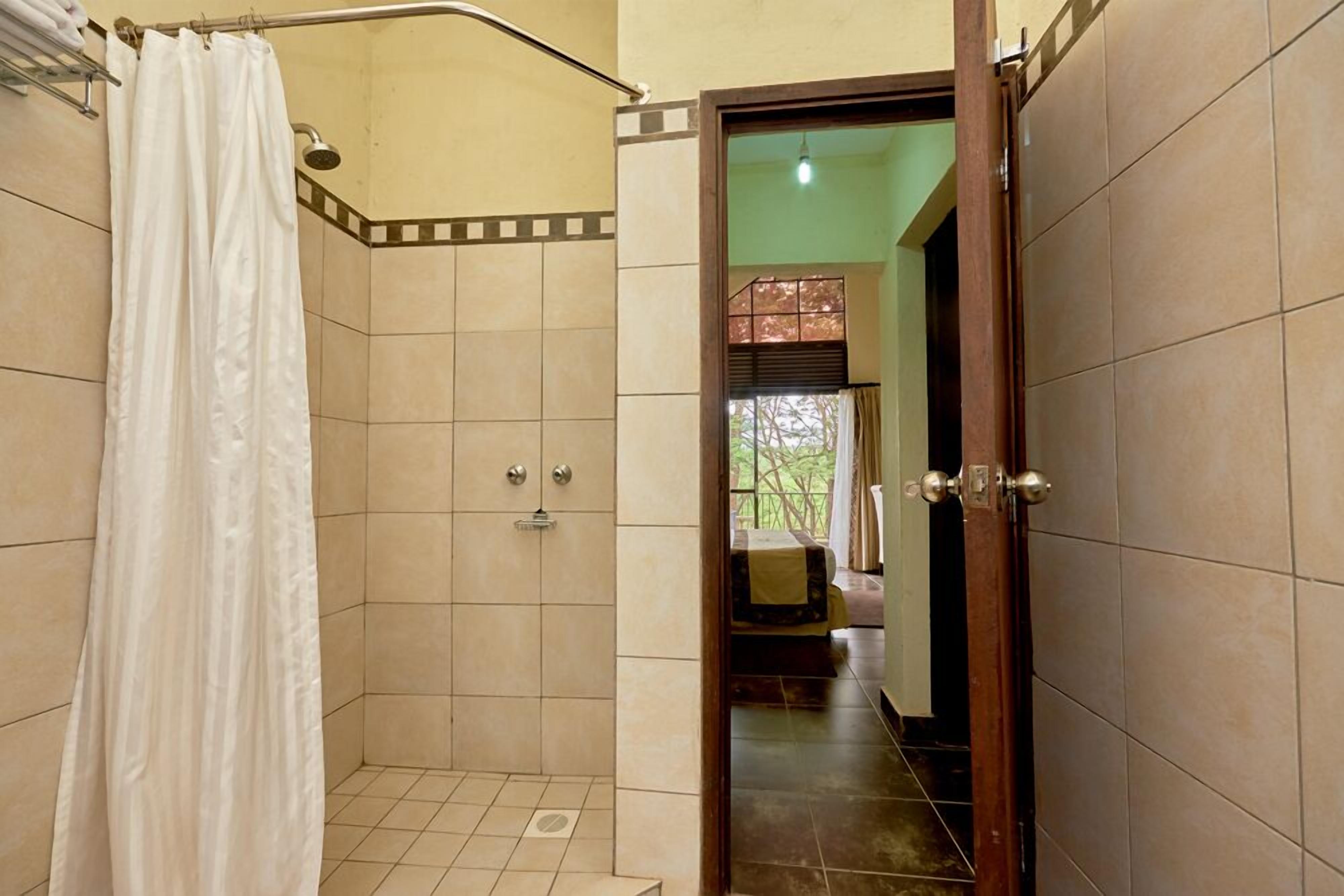 A shower area with a white curtain, tiled walls, and a door leading to a room with a bed and a view of trees.