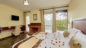 Habitación de hotel con cama, escritorio, silla, televisor y balcón con vistas a la vegetación.
