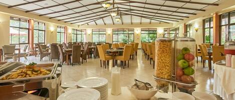 A spacious dining area with high ceilings, large windows, and a variety of food service stations including a fruit display and a buffet.
