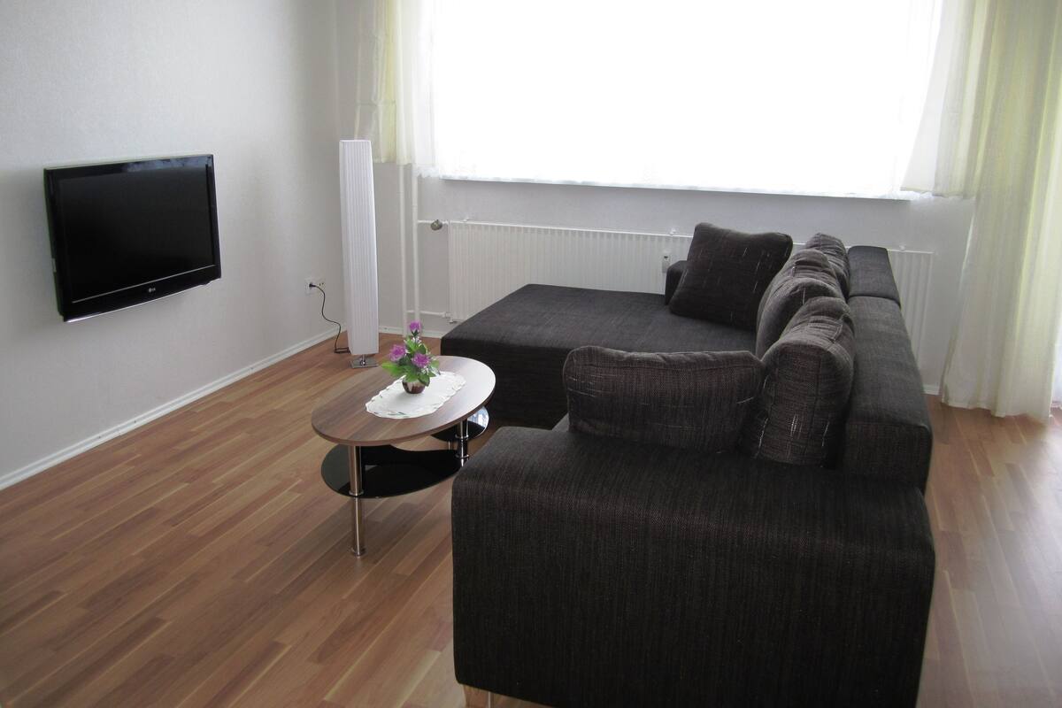Een moderne woonkamer met een donkergrijze bank, een flatscreentelevisie aan de muur en een kleine ronde tafel met een bloemenvaas.